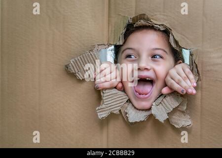 Fille souriante avec sa bouche ouverte derrière un carton. La fille regarde à travers un grand trou dans le carton et avec ses mains sur le bord du h Banque D'Images