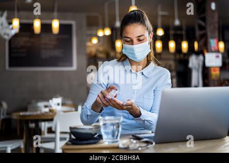 Portrait d'une jeune femme avec masque chirurgical en utilisant du gel désinfectant pour les mains au restaurant. Antiseptique, virus pandémique Corona, concept d'hygiène et de soins de santé. Banque D'Images