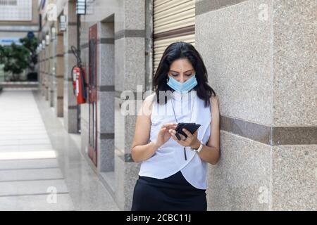 Une femme indienne portant un masque de protection Covid-19 regarde son téléphone dans un cadre urbain d'entreprise. Femme d'entreprise en tenue habillée élégante Banque D'Images