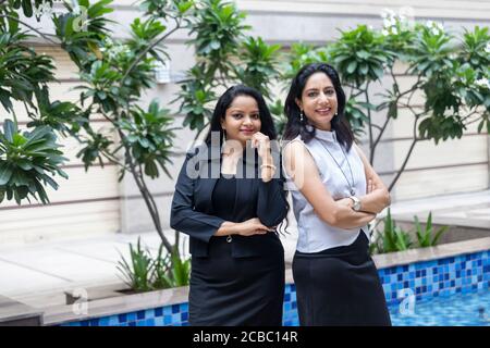 Deux femmes indiennes intelligentes debout côte à côte avec leurs armes croisées dans un cadre urbain d'entreprise. Banque D'Images