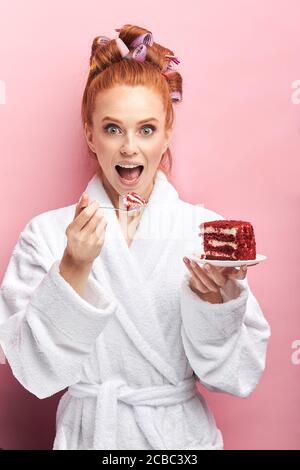 Jeune femme à tête rouge portant un peignoir, curlers après la douche mangeant un gâteau sucré et savoureux. Fille émotive posant avec le gâteau isolé sur fond rose Banque D'Images