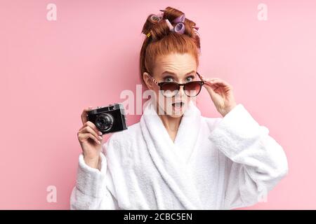 REDHEAD femme attrayante portant des lunettes de soleil, peignoir et curlers est surpris debout isolé sur fond rose, tenant photo appareil. Banque D'Images