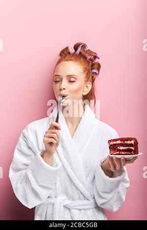 Jeune femme à tête rouge portant un peignoir, curlers après la douche mangeant un gâteau sucré et savoureux avec une cuillère. Fille émotive posant avec le gâteau isolé sur le dos rose Banque D'Images