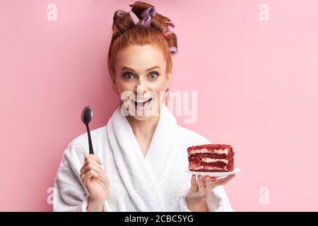 Jeune femme à tête rouge portant un peignoir, curlers après la douche mangeant un gâteau sucré et savoureux. Dans une cuillère à main, dans un autre gâteau rouge. Fille émotive posant avec Banque D'Images