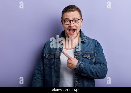 Jeune homme joyeux et joyeux, portant une veste en denim à large bouche ouverte, pointant de la main et du doigt sur le côté de la caméra.publicité, Banque D'Images
