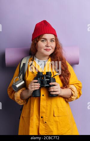 agréable femme belle apparence dans des vêtements élégants tenant ses jumelles et posant à l'appareil photo. portrait gros plan, isolé bleu arrière-plan studio sho Banque D'Images