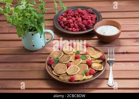 Céréales pancakes et framboises fraîches mûres rouges dans une assiette brune. Un bouquet de Melissa dans une tasse sur une table en bois. Délicieux petit déjeuner d'été Banque D'Images