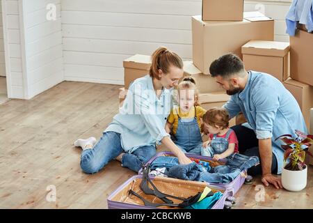 Famille caucasienne, homme, femme et deux filles, s'asseoir sur le sol, sourire dans le salon nouvelle maison. L'homme et la femme déballent la valise et les enfants aident. Derrière le Banque D'Images