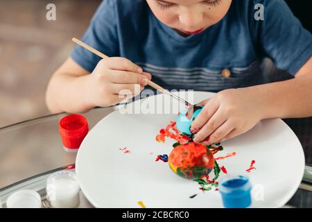 Petit enfant mâle étonnant assis à table et apprenant à peindre les oeufs de Pâques, se concentrer sur les mains. Étude de créativité des enfants, concept de divertissement Banque D'Images