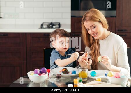 Souriant agréable jeune blonde femme et son enfant mignon se préparer pour Pâques en peignant les oeufs à la maison. Banque D'Images