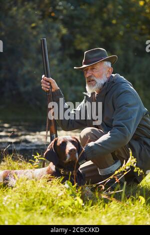 Chasseur de bonne qualité senior avec son chien de garde à la chasse sur les canards sauvages. Assis près du lac, portant un chapeau, tenant un fusil Banque D'Images