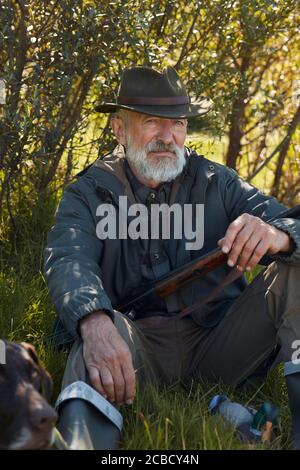 Chasseur senior avec arme à feu s'assoir sur l'herbe avec chien après chasse Banque D'Images