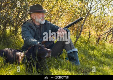 Chasseur senior avec arme à feu s'assoir sur l'herbe avec chien après chasse Banque D'Images