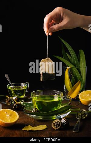 Personne met un sac d'herbes dans une tasse de verre de thé vert chaud sur une table en bois avec des feuilles d'eucalyptus, des feuilles de Laurier, des épices telles que l'origan et les citrons. N/a Banque D'Images