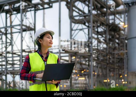 Femme asiatique ingénieur pétrochimique travaillant la nuit avec un ordinateur portable dans l'usine de raffinage de pétrole et de gaz la nuit pour la sécurité des inspecteurs Banque D'Images