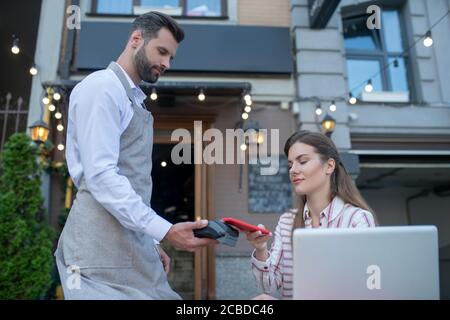 Serveur tenant la machine de paiement, femme brun payant facture avec smartphone Banque D'Images