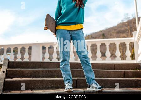 Une jeune adolescente tenant un skateboard derrière son dos est debout sur les marches. Vue de dessous. Gros plan. Concept de style de vie sportif et de culture de rue. Banque D'Images