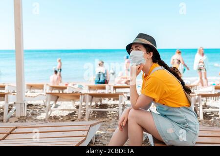 Portrait d'une femme dans un masque médical s'ennuie sur une chaise longue, son menton a appuyé sur sa main. En arrière-plan, la plage et la mer. Le concept Banque D'Images