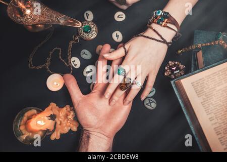 Astrologie et ésotéricisme. Un joueur de fortune lit la paume d'un homme. Sur fond noir se trouvent des runes, une bougie, des amulettes et un livre. La vue du dessus. Banque D'Images