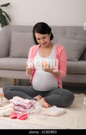 belle femme asiatique enceinte utilisant la liste d'écriture de stylo sur le bloc-notes emballage et préparation des vêtements de bébé dans le panier Banque D'Images