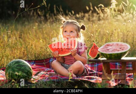 une petite fille drôle sur un pique-nique d'été mange un rouge mûr délicieux pastèque juteuse. Banque D'Images