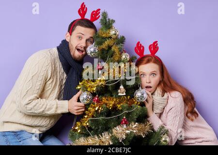 Belle jeune fille aux cheveux longs rouges, vêtue d'un chandail beige chaud, essayant de se cacher sous un arbre de Noël décoré, regardant droit avec grand ouvert Banque D'Images
