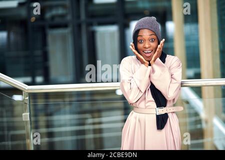 Joyeuse surprise jeune musulmane d'origine africaine vêtue robe longue rose et foulard noir posant dans un cadre urbain sourire à l'appareil photo Banque D'Images