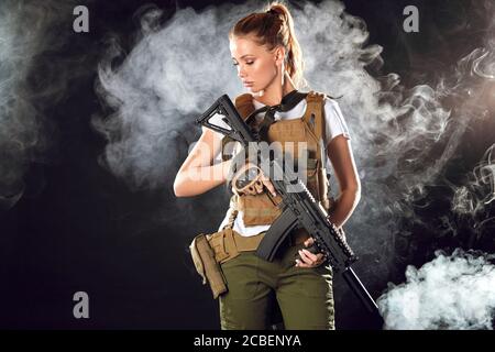 Belle femme caucasienne entrepreneur militaire privé avec de longs cheveux blonds, vêtu d'un uniforme tactique, armé de volants posant isolés en studio Banque D'Images