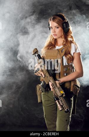 Femme soldat en uniforme de camouflage militaire protégée par un casque, une armure corporelle, tenant une carabine d'assaut debout sur un fond sombre et fumé Banque D'Images