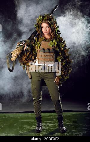 Une femme camouflée sniper en costume de ghillie posant avec le fusil dans la nuit brumeuse. Concept d'armes spéciales et de femme. Banque D'Images