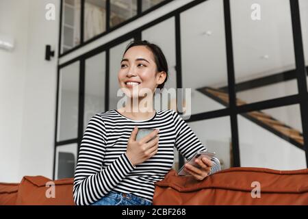 Jolie jeune femme asiatique souriante portant des vêtements décontractés se détendant sur un canapé en cuir à la maison, à l'aide d'un téléphone portable Banque D'Images