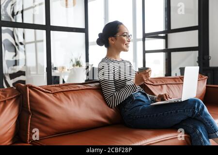 Jolie jeune femme asiatique souriante vêtue de vêtements décontractés se détendant sur un canapé en cuir à la maison, travaillant sur un ordinateur portable Banque D'Images