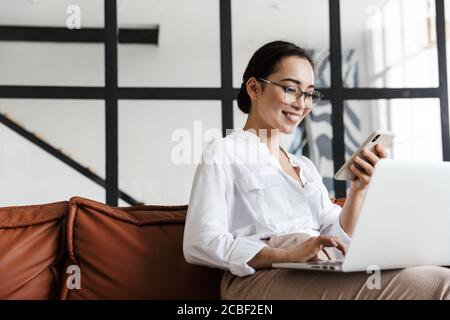 Jolie jeune femme d'affaires asiatique souriante se détendant sur un canapé en cuir à la maison, travaillant sur un ordinateur portable, tenant un téléphone portable Banque D'Images