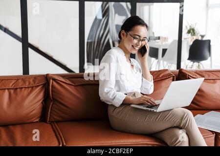 Belle jeune femme d'affaires asiatique souriante se détendant sur un canapé en cuir à la maison, travaillant sur un ordinateur portable, parlant sur un téléphone portable Banque D'Images