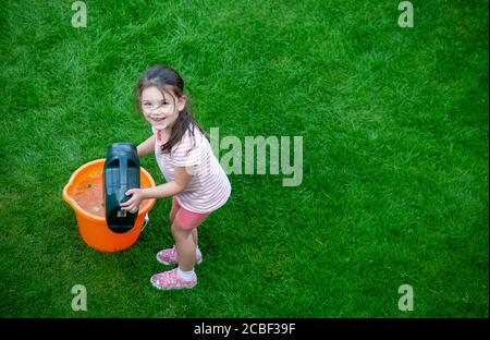 Fille de 4 ans, portant des vêtements d'été, versant de l'eau d'une arrosoir verte dans un seau orange dans un jardin anglais. Lancashire Royaume-Uni Banque D'Images