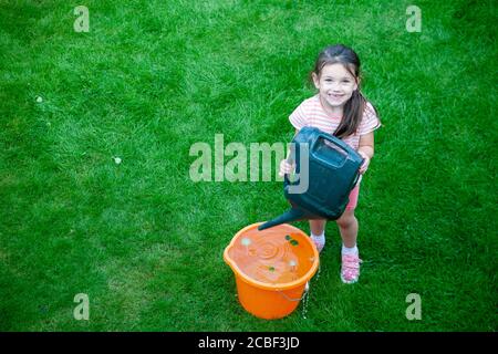 Fille de 4 ans, portant des vêtements d'été, versant de l'eau d'une arrosoir verte dans un seau orange dans un jardin anglais. Lancashire Royaume-Uni Banque D'Images