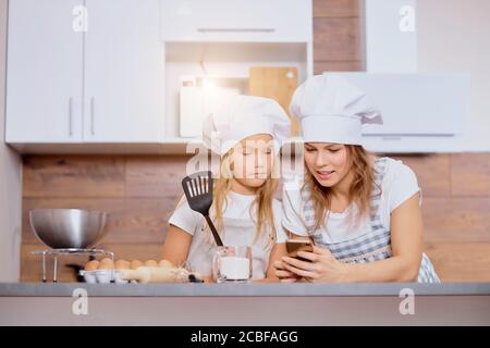 la mère caucasienne et la fille d'enfant en casquettes et tabliers regardent l'écran de smartphone regarder la recette de gâteau délicieuse, allant cuisiner ensemble Banque D'Images