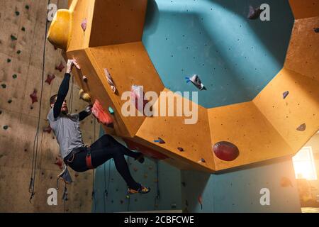 Vue à angle bas d'un sportif handicapé, grimpeur puissant se déplaçant sur un mur de rochers escarpé, s'entraîner dans un grand centre de bloc intérieur coloré Banque D'Images