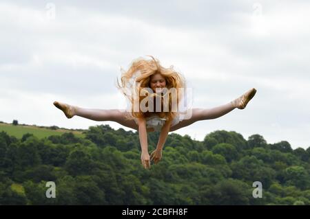 Une jeune ballerine portant un tutu blanc faisant un saut en deux dans l'air, ses longs cheveux blonds survolant. Banque D'Images