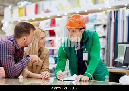 les jeunes couples mariés caucasiens veulent obtenir des produits de base sur le marché, un employé d'entrepôt amical les aider avec le choix et les conseils, ils ont la conversation Banque D'Images