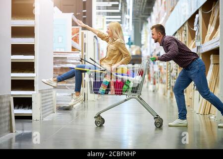 couple heureux marié shopping dans le magasin, femme assis sur le chariot, mari gai roule son dans hypermarché. concept de shopping Banque D'Images