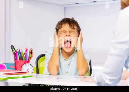 Garçon avec le trouble de l'autisme cri et les oreilles proches dans la douleur pendant le traitement de développement de l'ABA assis par la table en classe, problème sensoriel Banque D'Images