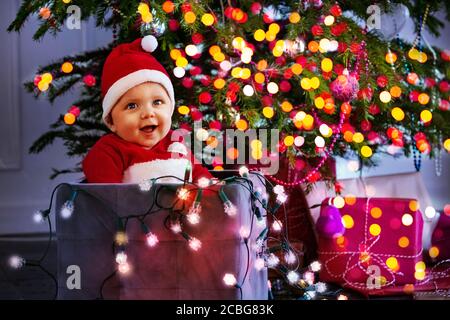 Petit garçon mignon dans un chapeau du Père Noël tenant illuminé Lampes placées dans une boîte sous l'arbre de Noël avec des lumières floues Banque D'Images