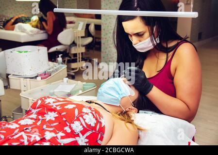 Jeune femme brunette aux cheveux longs mettant de fausses cils sur la dame en salon de beauté avec des masques, nouvelle normale Banque D'Images