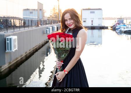 Portrait d'une jeune femme attrayante avec des cheveux bruns en petit robe noire souriant et tenant un bouquet de roses rouges en arrière-plan du restaurant à Banque D'Images