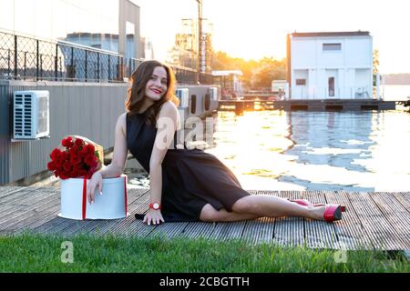 Jolie fille d'anniversaire avec des cheveux bruns dans une petite robe noire assise sur le sentier en bois au coucher du soleil. Boîte ronde avec gâteau d'anniversaire enveloppé de côtes rouges Banque D'Images