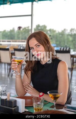 Portrait d'une jeune jolie femme assise à une table dans le restaurant tenant le verre à cocktail regardant l'appareil photo. Concept d'idée d'anniversaire Banque D'Images