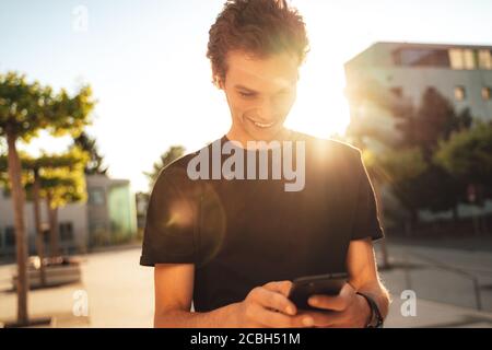 Jeune homme souriant lisant un message texte sur un smartphone en ville par beau temps Banque D'Images