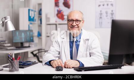 Médecin mature portant des lunettes et souriant dans le cabinet de l'hôpital assis au bureau. Banque D'Images