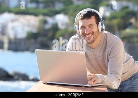 Un homme heureux avec un casque et un ordinateur portable qui se pose en regardant l'appareil photo sur la plage Banque D'Images